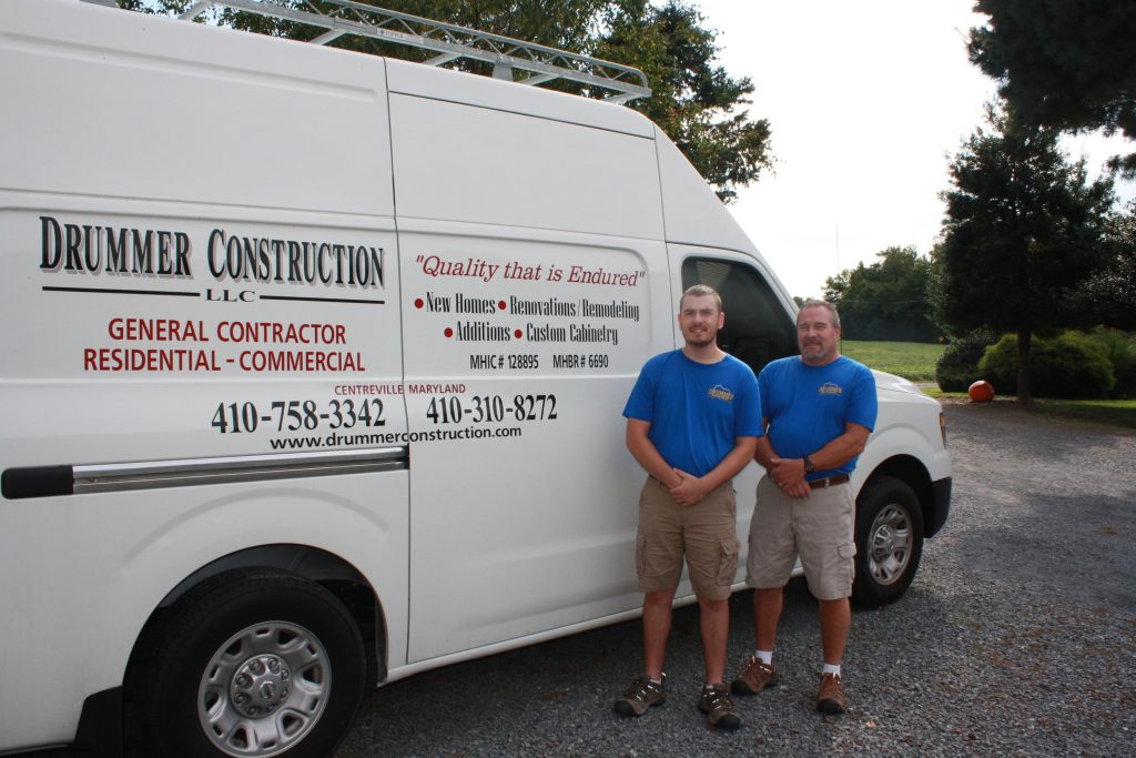 Two individuals standing beside a white van bearing the Drummer Construction LLC logo and contact information.
