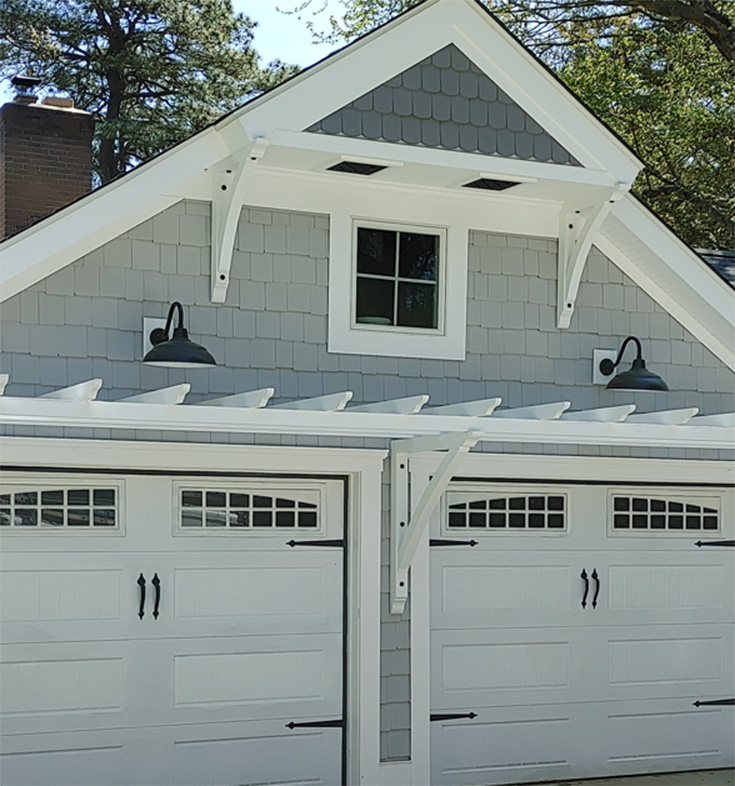 A garage with white doors and a gable above featuring a window and outdoor wall lanterns.