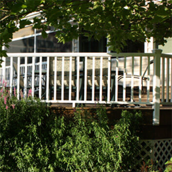 A wooden deck with white railings, outdoor chairs, and surrounded by greenery and flowering shrubs in a backyard.
